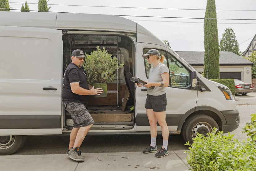 Two R&M crew members unloading a container shrub from a delivery van at a residential curb.